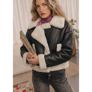 May include: A black jacket with a white faux fur lining, wide lapels, and cuffs. The jacket features a front zipper closure and is worn with a purple patterned neck scarf and dark gray denim jeans. The jacket has a cropped fit and long sleeves.