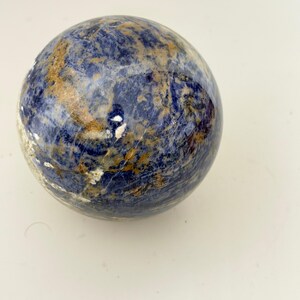 May include: A polished spherical stone featuring a marbled pattern of deep royal blue, white, and earthy tan veins. The surface is smooth and reflective, showcasing natural mineral inclusions and varied color transitions across the entire round form.