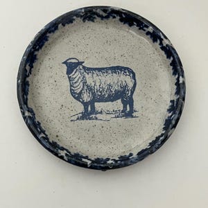 May include: A round ceramic plate with a speckled light gray base and a dark blue sheep illustration in the center. The rim features a dark blue mottled pattern. The plate has a shallow basin and a raised edge, showcasing a rustic, speckled finish.