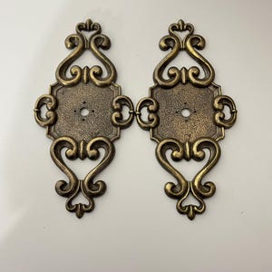 May include: Two matching antique brass-toned metal plates featuring an elongated, symmetrical scrollwork design. Each piece has a central rectangular section with a textured surface and a small circular hole, flanked by ornate, curved metal flourishes.