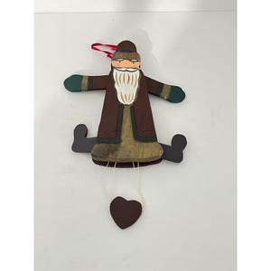 May include: A wooden hanging ornament shaped like a bearded figure wearing a brown coat and hat, green mittens, and black boots. The figure has a white beard, pink cheeks, and a dark brown heart-shaped weight suspended by white strings below.