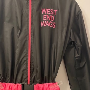 Könnte beinhalten: Schwarzer Nylon-Overall mit einem rosafarbenen Reißverschluss und Gürtel. Der Overall hat den Text "WEST END WAGS" in rosa auf der Vorderseite gestickt.