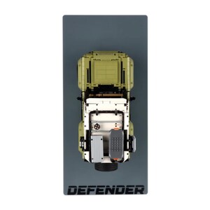 May include: Overhead shot of an olive green, white, and grey model Defender SUV. The model is on a grey rectangular base with the word "DEFENDER" in black. The model has detailed features.
