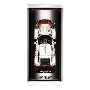 May include: A white Lego Porsche RSR race car model displayed in a clear acrylic display case with a black background. The car is in a top-down view and the text "RSR PORSCHE" is printed on the base.