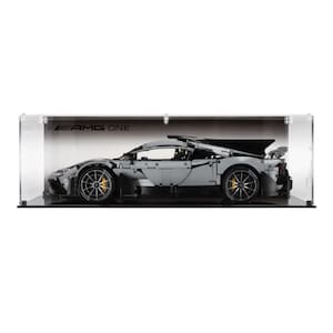 May include: A grey and black model sports car with yellow detailing, presented in a clear acrylic case. The car has detailed features, including visible wheels and a rear spoiler. The text "AMG ONE" is visible on the case.