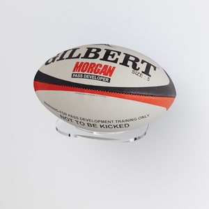 May include: White and red rugby ball with black trim. The ball is labelled "Gilbert Morgan Pass Developer Size: 5" and has a warning that reads "Warning for pass development training only. Not to be kicked."