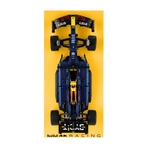 May include: A navy blue and yellow race car model, viewed from above, set against a yellow backdrop. The model features black wheels and the text "BYBIT" on the front and rear wings. The words "Red Bull Racing" are at the bottom.
