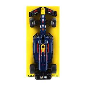 May include: A dark blue and yellow Formula 1 model car, viewed from above. The model displays Red Bull branding and sponsor logos. The car is positioned on a bright yellow surface.
