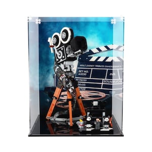 May include: A clear acrylic display case with a black base, showcasing a LEGO® model of a vintage movie camera with a tripod. The camera is made of black, white, and orange LEGO® bricks. The background of the display case features a blue and white backdrop with a film reel and a clapperboard. The clapperboard has the text "WALT DISNEY TRIBUTE CAMERA" and "SET NO. 43230".