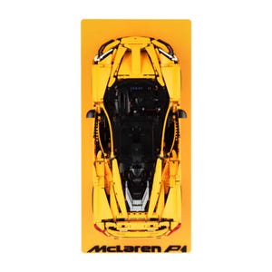 May include: A yellow McLaren P1 model car, viewed from above, set against a yellow backdrop. The black interior and the McLaren P1 logo are clearly visible. The model is a detailed replica of the sports car, showcasing its design.