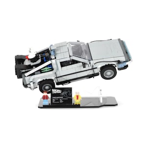 May include: A grey DeLorean time machine model from the Back to the Future film, with a black base and two mini-figures. The model is elevated, showcasing the car's details, including wheels and engine components. The base has the text "BACK TO FUTURE" and "TIME MACHINE".