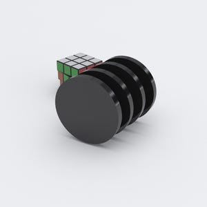 May include: Five black, circular objects stacked together with a glossy finish. A Rubik's Cube with red, white, green, blue, orange, and yellow squares is in the background. The objects are on a white surface.