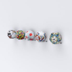 May include: Five footballs are displayed on a white wall mounted rack. The balls are all different designs and colours. The balls are all in focus and the rack is in the background.