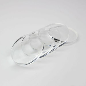 May include: Four clear, circular coasters are arranged in a slightly curved line. The coasters are made of transparent material, allowing light to pass through. The edges of the coasters are smooth and rounded. The background is plain white.