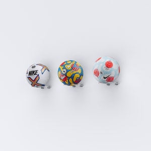 May include: Three footballs with different designs are displayed on a white wall. The footballs are mounted on a wall with a metal bracket. The footballs are all white with different coloured designs. One football has a blue, red, and yellow design. Another football has a blue, red, and white design. The third football has a blue, red, and white design with the word "NIKE" on it.