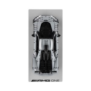 May include: Overhead shot of a light grey model car with black detailing, on a rectangular grey base. The car's design is intricate, showcasing its engine and chassis. The base features the text "AMG ONE" in black.
