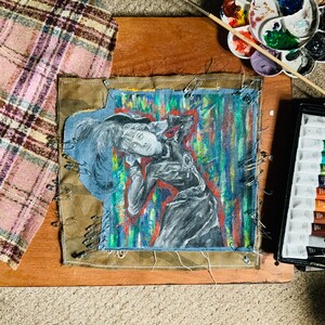 Könnte beinhalten: Eine Nahaufnahme eines Stoffkunstwerks mit einer Zeichnung einer Person mit mehreren Augen auf blauem Hintergrund. Der Stoff ist mit Sicherheitsnadeln an einem Holzbrett befestigt. Das Kunstwerk ist von bunter Farbe und einer Farbpalette umgeben.