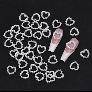 May include: A collection of white pearl heart-shaped nail charms. The charms are made of small white pearls arranged in a heart shape. There are several charms scattered on a black surface and two nail charms are on a fingernail.