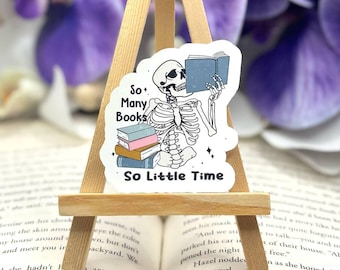 Może przedstawiać: Naklejka przedstawiająca szkielet trzymający książkę z tekstem "So Many Books, So Little Time". Naklejka znajduje się na małym drewnianym sztalugach i jest otoczona fioletowymi kwiatami.