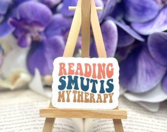 May include: A white sticker with a colorful design that reads "Reading Smut is My Therapy". The sticker is on a small wooden easel.