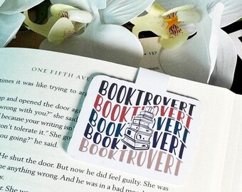 Op de afbeelding: Een witte bladwijzer met de tekst "BOOKTROVERT" die herhaald wordt in een ontwerp van een stapel boeken. De tekst is in blauw, rood en zwart. De bladwijzer ligt op een boek.
