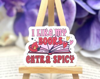 May include: A sticker with the text "I LIKE MY BOOKS EXTRA SPICY" in pink and red, featuring an open book with hearts and a chili pepper. The sticker is displayed on a small wooden easel, with a blurred purple floral background.