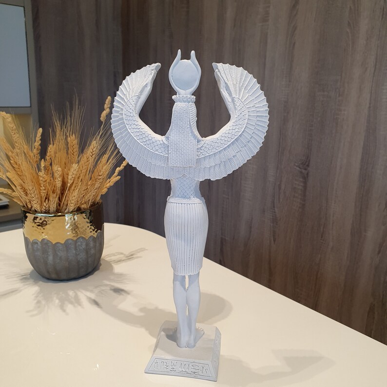 May include: A white statue of an Egyptian goddess with outstretched wings and a headdress. The statue is standing on a rectangular base with hieroglyphs. The statue is on a white table with a decorative vase of wheat in the background.