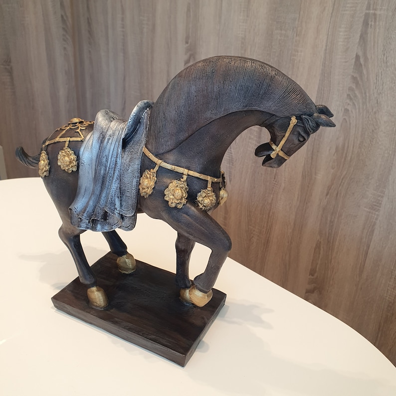 May include: A black horse figurine with gold accents and a blue fabric saddle. The horse is standing on a wooden base.