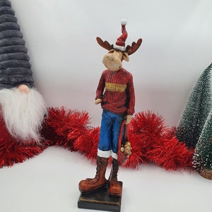 May include: A red and white Christmas reindeer figurine with a red sweater, blue jeans, and a bag of presents. The reindeer is standing on a brown base.