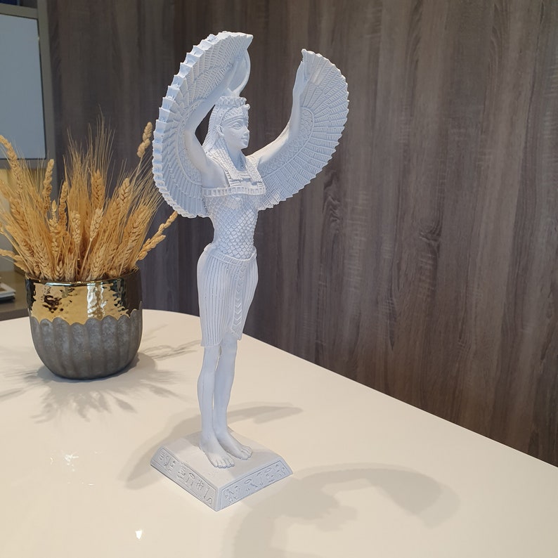 May include: A white statue of an Egyptian goddess with outstretched wings. The figure is standing on a rectangular base with hieroglyphs. The statue is on a white surface, with a decorative vase of wheat stalks in the background.