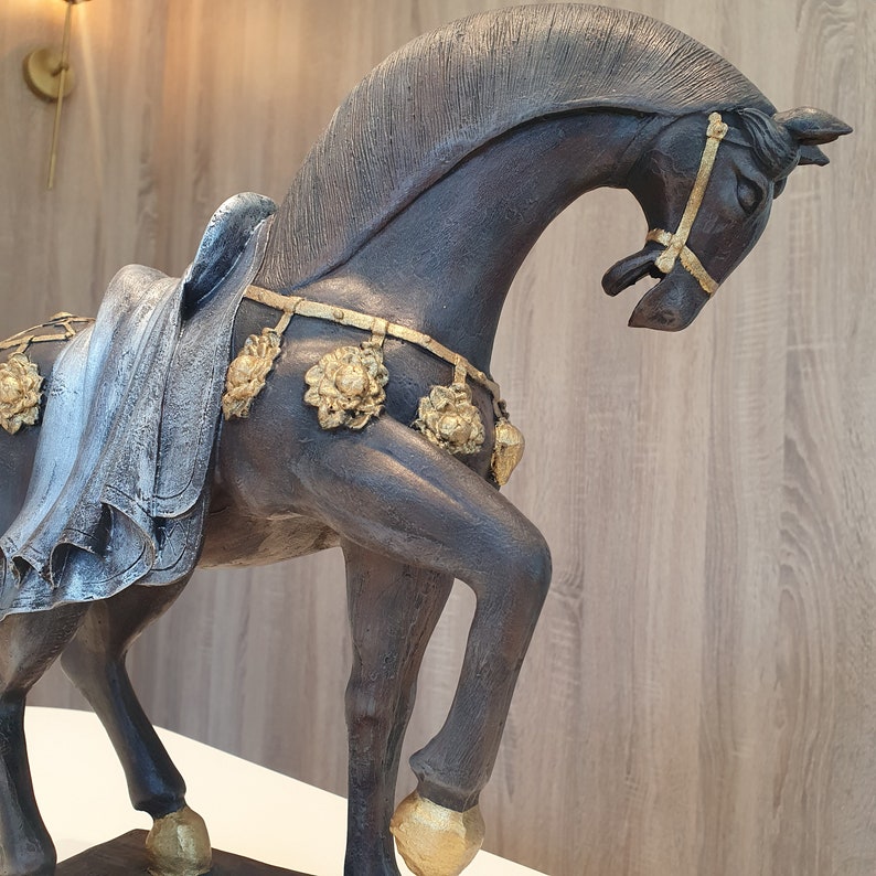 May include: A dark brown horse figurine with gold accents. The horse is wearing a decorative harness with gold flowers and is standing on a wooden base.