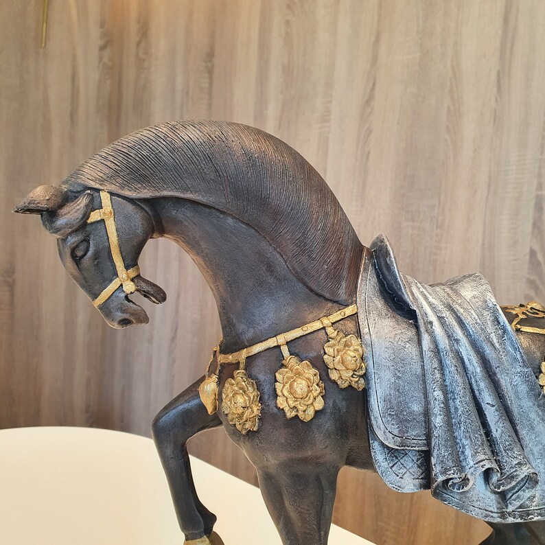 May include: A black horse figurine with gold accents and a draped fabric saddle. The horse has a detailed head and mane, and the saddle is decorated with floral patterns.