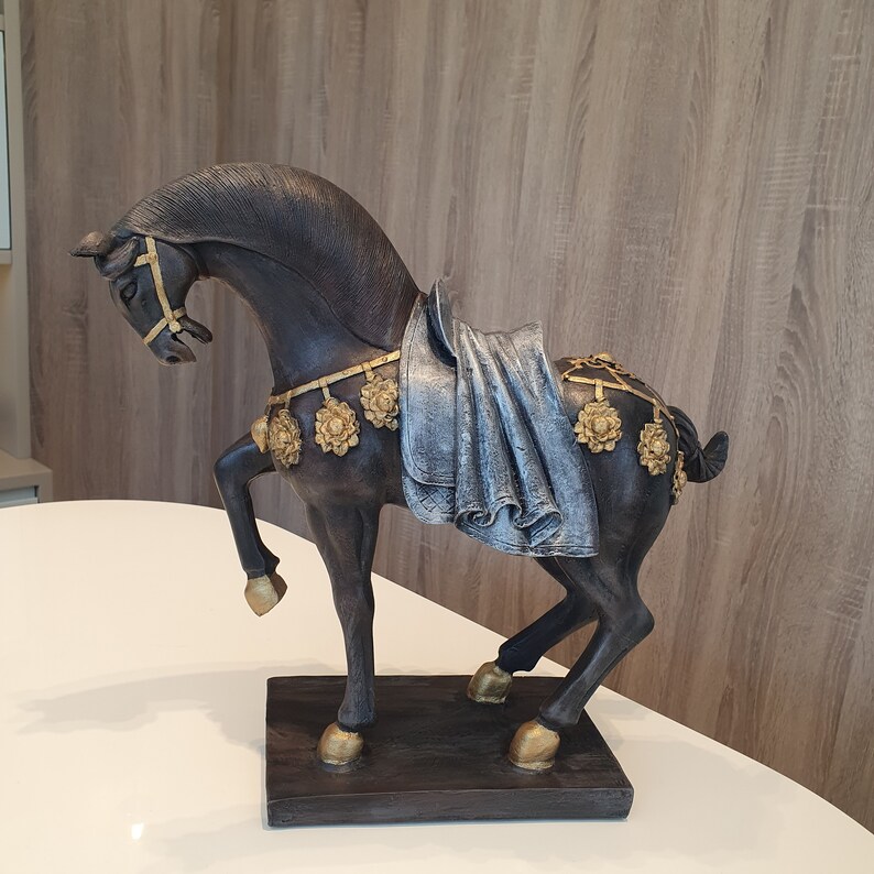 May include: A black horse figurine with gold accents and a blue and silver fabric draped over its back. The horse is standing on a black base.