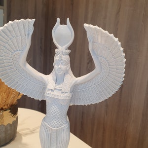 May include: A white statue of an Egyptian goddess with outstretched wings and a headdress. The statue has intricate details, including the goddess's face, clothing, and the feathers of her wings. The statue is on a white surface.