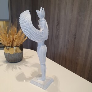 May include: A white statue of a figure with large, detailed wings, standing on a rectangular base with hieroglyphs. The figure has a headdress and is posed in a side profile. The statue is on a white surface.