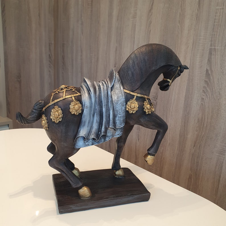 May include: A dark brown horse figurine with a blue and silver fabric draped over its back. The horse has gold accents and is standing on a dark brown wooden base.