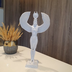 May include: A white statue of an Egyptian goddess with outstretched wings and a headdress. The figure stands on a rectangular base with hieroglyphs. The statue is on a white surface, with a decorative vase of wheat stalks in the background.