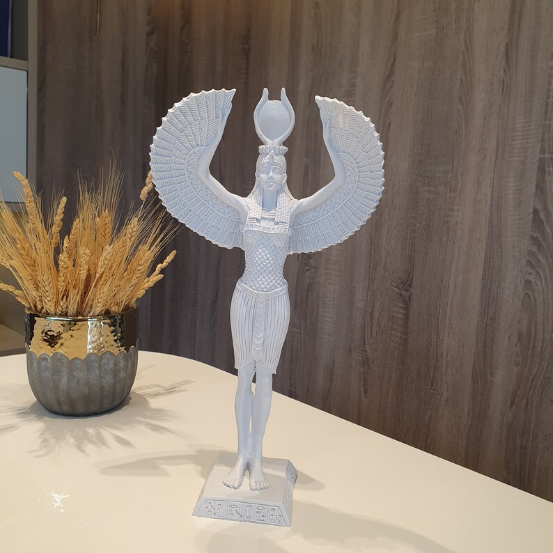 May include: A white statue of an Egyptian goddess with outstretched wings and a headdress. The figure stands on a rectangular base with hieroglyphs. The statue is on a white surface, with a decorative vase of wheat stalks in the background.