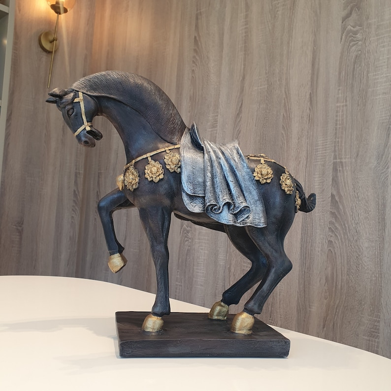 May include: A black horse figurine with gold accents and a silver fabric draped over its back. The horse is standing on a black base.