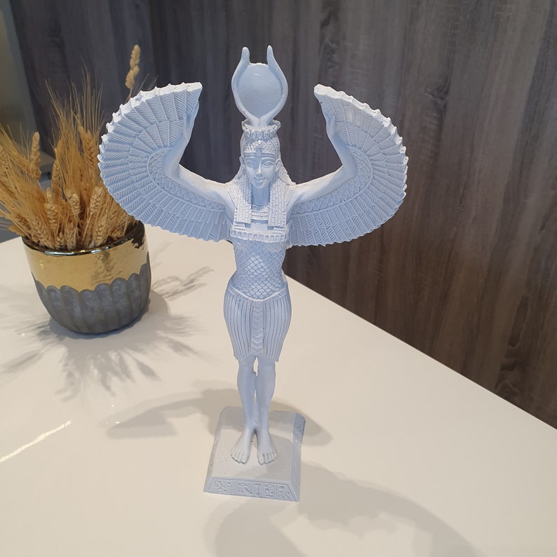 May include: A light blue statue of an Egyptian goddess with outstretched wings and a headdress. The statue is standing on a rectangular base. The statue is on a white surface.