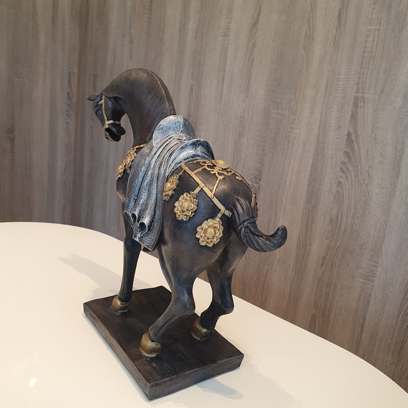 May include: Black horse figurine with gold accents and a blue and white fabric saddle. The horse is standing on a black base.