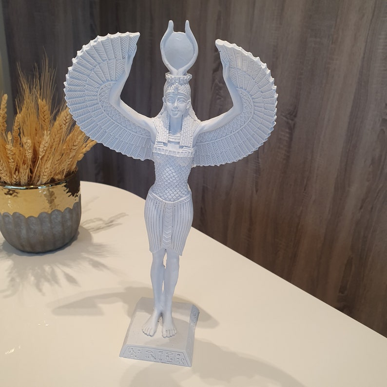 May include: A light blue statue of an Egyptian goddess with outstretched wings and a headdress. The statue is standing on a rectangular base with hieroglyphs. The statue is on a white surface.