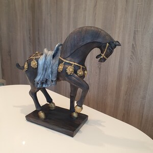 May include: A black horse figurine with gold accents and a blue and silver fabric draped over its back. The horse is standing on a dark brown base.