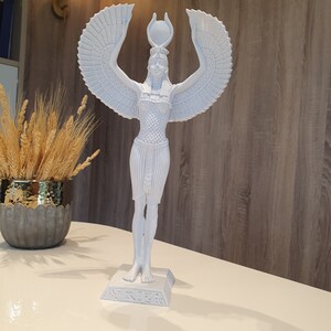 May include: A white statue of an Egyptian goddess with outstretched arms and large, detailed wings. The statue has a detailed headdress and is standing on a rectangular base with hieroglyphs. The statue is approximately 12 inches tall.