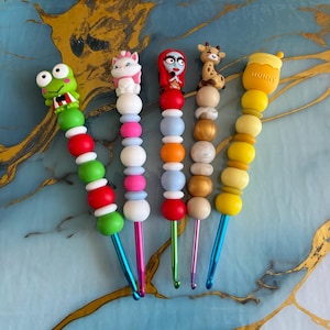 Puede incluir: Cinco ganchillos con adornos de silicona en la parte superior que representan una rana verde, un gato blanco, un personaje pelirrojo, una jirafa y un tarro de miel amarillo con la palabra HUNNY. Cada mango tiene cuentas de silicona de colores.