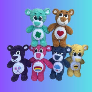 Può includere: Sette orsacchiotti di peluche all'uncinetto in vari colori, ognuno con un simbolo diverso sul petto. Gli orsi sono in piedi su uno sfondo sfumato blu e viola.