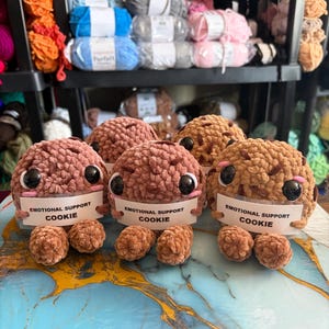Peut inclure: Des jouets en forme de cookies en peluche faits à la main, de différentes nuances de marron. Chaque cookie a de grands yeux noirs et une pancarte indiquant "EMOTIONAL SUPPORT COOKIE". Les cookies sont exposés sur une surface bleue et blanche, avec des étagères de laine en arrière-plan.