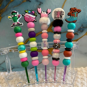 Puede incluir: Cinco ganchillos con adornos decorativos y cuentas de colores. Los adornos incluyen un pepinillo con el texto 'let's pickle!', un ovillo de lana con 'CROCHETALLDAY', un cangrejo rosa, un cartel circular 'CRAZY CUP LADY' y una bota vaquera.