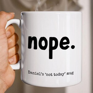 May include: A white ceramic mug featuring the word nope. printed in large black sans-serif letters. Below the main text, smaller black typewriter-style text reads Daniel's 'not today' mug. The mug has a curved handle on the left side.