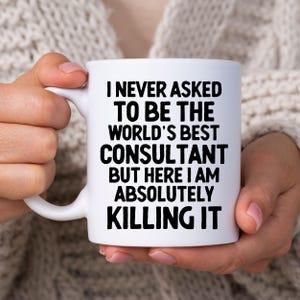 Könnte beinhalten: Ein weißer Keramikbecher mit fettgedrucktem schwarzem Text, der lautet: I NEVER ASKED TO BE THE WORLD'S BEST CONSULTANT BUT HERE I AM ABSOLUTELY KILLING IT. Der Becher hat einen Standardhenkel auf der linken Seite und ein glattes, zylindrisches Design.